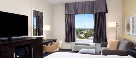 Habitación de hotel con una cama grande, un escritorio con televisor de pantalla plana, una silla, un sofá, una lámpara y vistas a la ciudad.