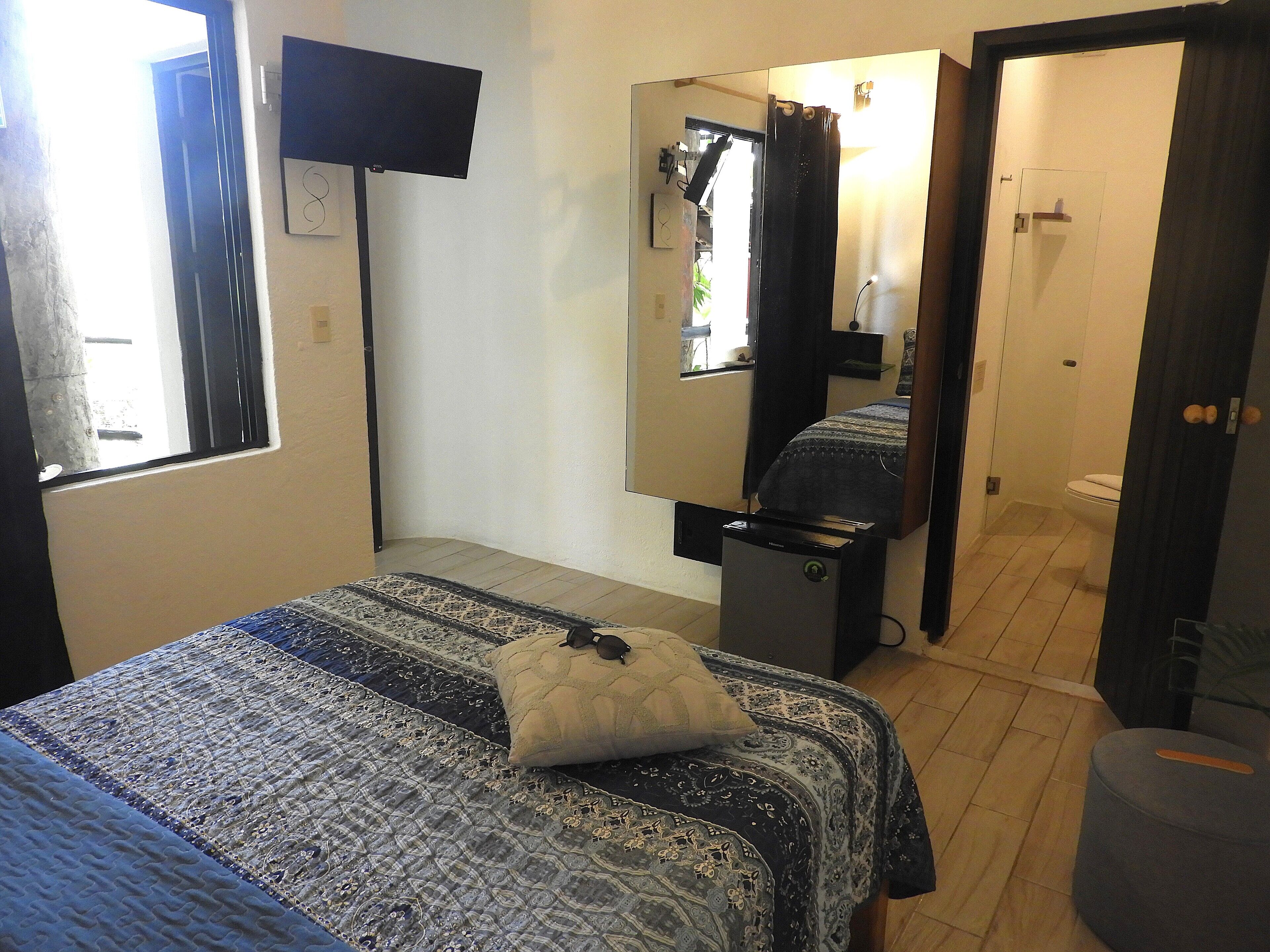 A bedroom with a bed, a bedside table, a mirror, a window, and a door leading to a bathroom.