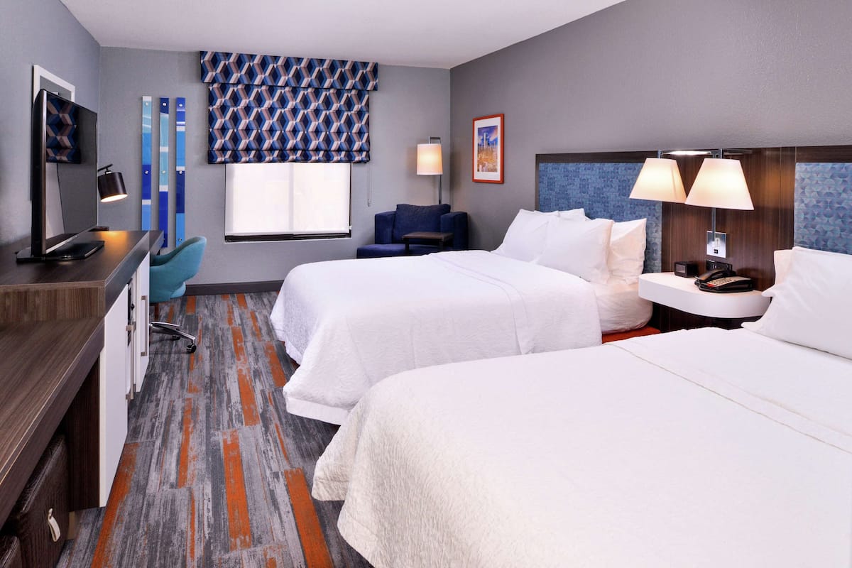 Habitación de hotel con dos camas, un escritorio con una computadora, una silla, una lámpara y una ventana con cortinas estampadas.