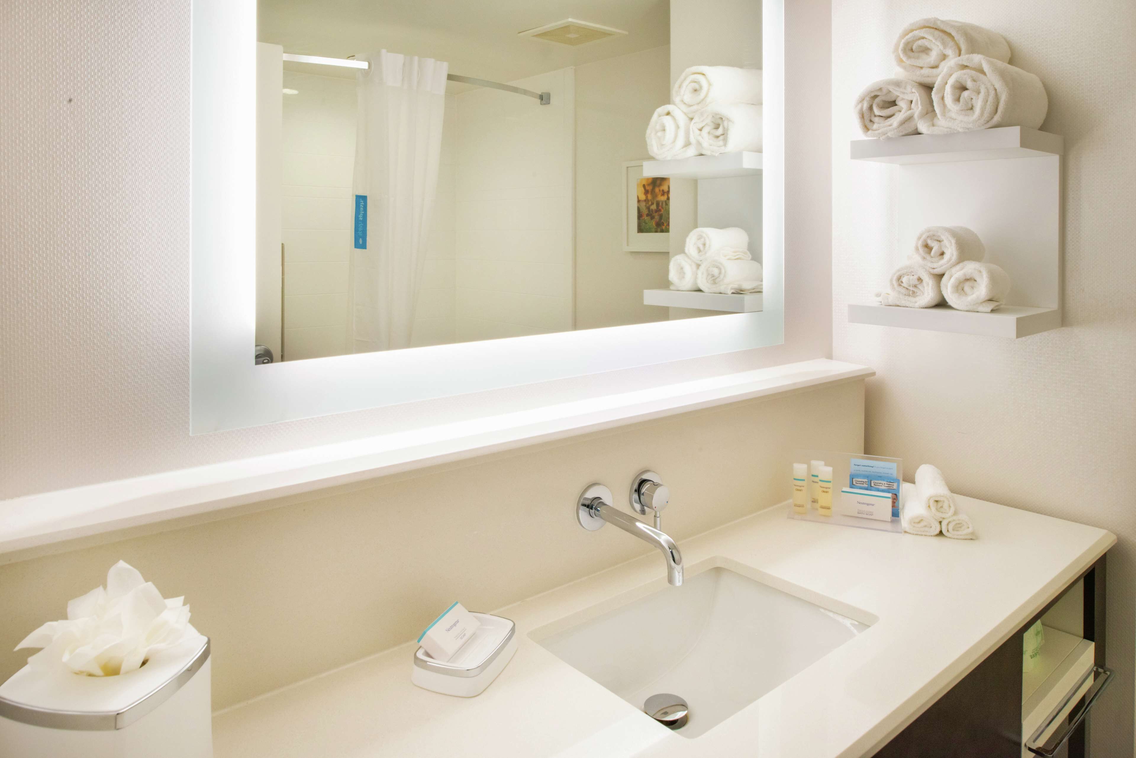 Une salle de bain d’hôtel avec un grand miroir, un lavabo avec un robinet et des étagères où sont pliés des serviettes.