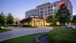 Ein Hampton Inn Hotel mit gut beleuchtetem Eingang, angelegten Grünflächen und mehreren Parkplätzen.