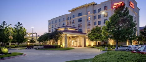 Un hotel Hampton Inn con una entrada bien iluminada, jardines ajardinados y varios espacios de estacionamiento.