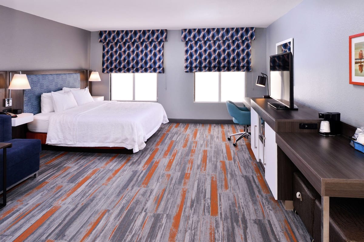 Habitación de hotel con cama, escritorio, silla y dos ventanas con cortinas estampadas.