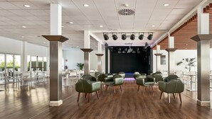 A spacious indoor area with a stage at the front, surrounded by rows of chairs and tables, and large windows on the sides. - Hotel Globales Cala´n Bosch (Ciutadella de Menorca)
