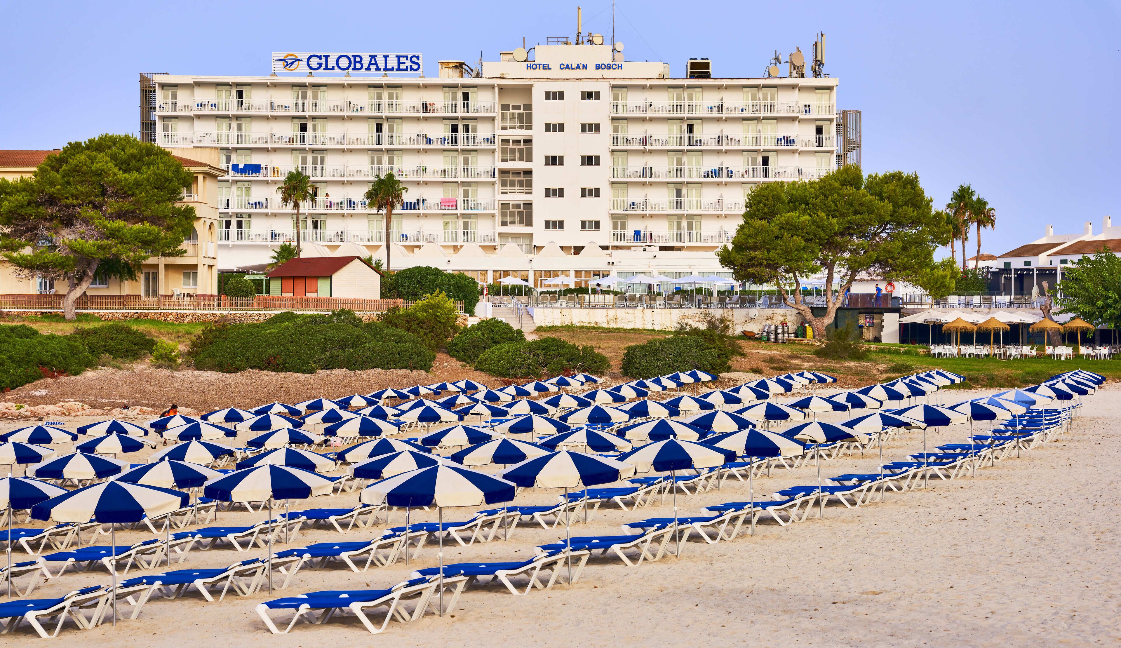Een hotel met meerdere verdiepingen, terras en een strand met rijen ligbedden en parasols.
