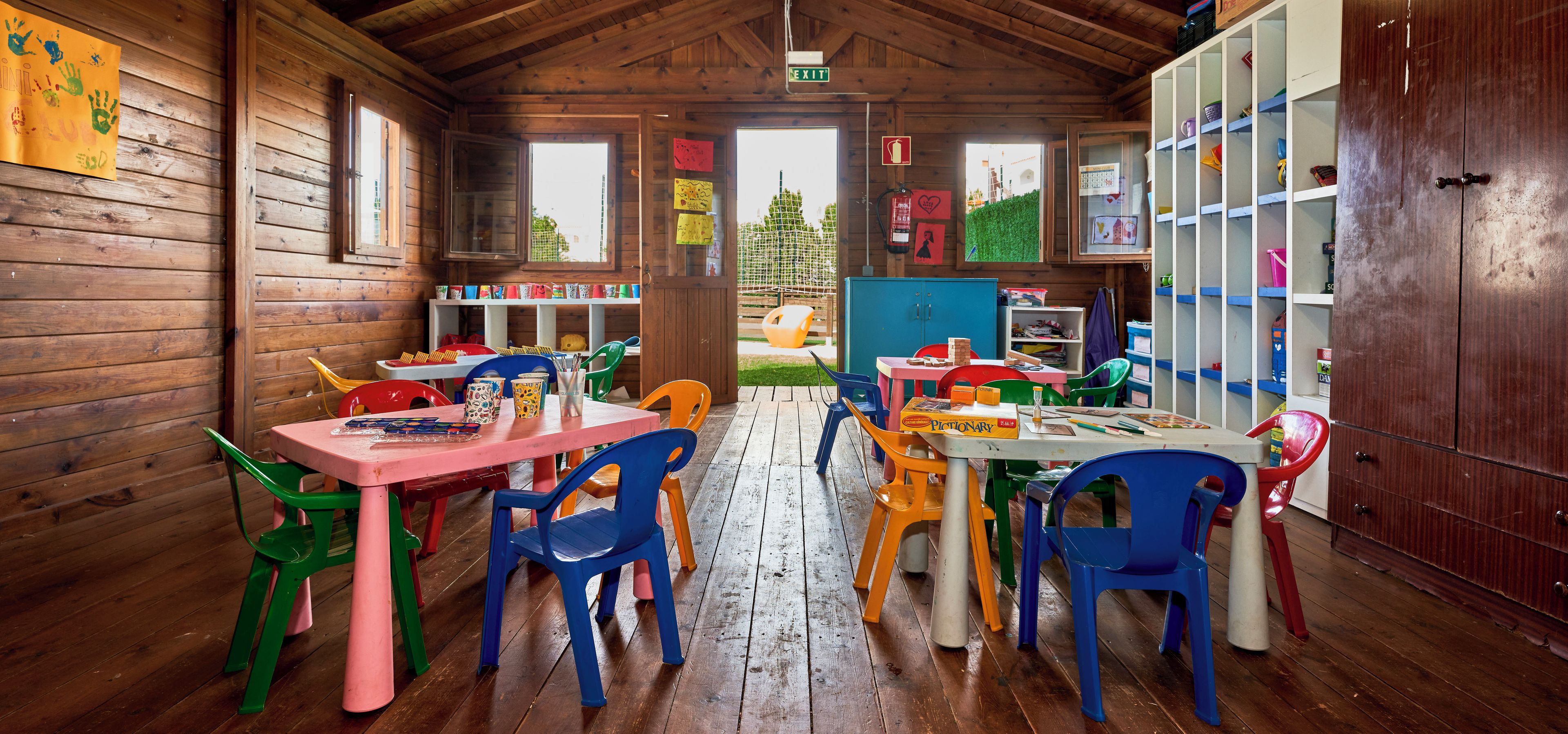 A wooden interior with multiple tables and colorful plastic chairs, a bookshelf, and an exit sign.