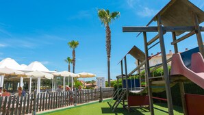A playground with slides and climbing structures, surrounded by palm trees and umbrellas. - Hotel Globales Cala´n Bosch (Ciutadella de Menorca)