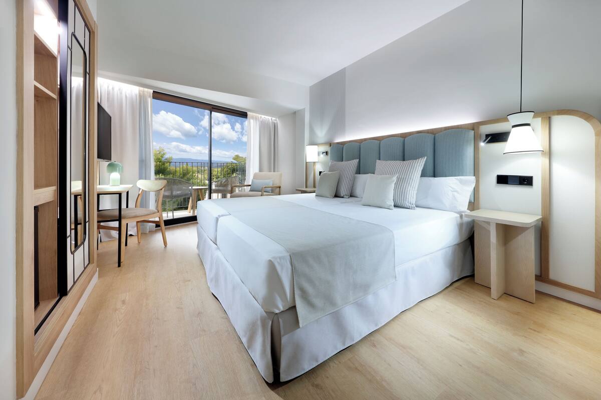 A modern hotel room with a large bed, a desk, a chair, and a balcony with a view.