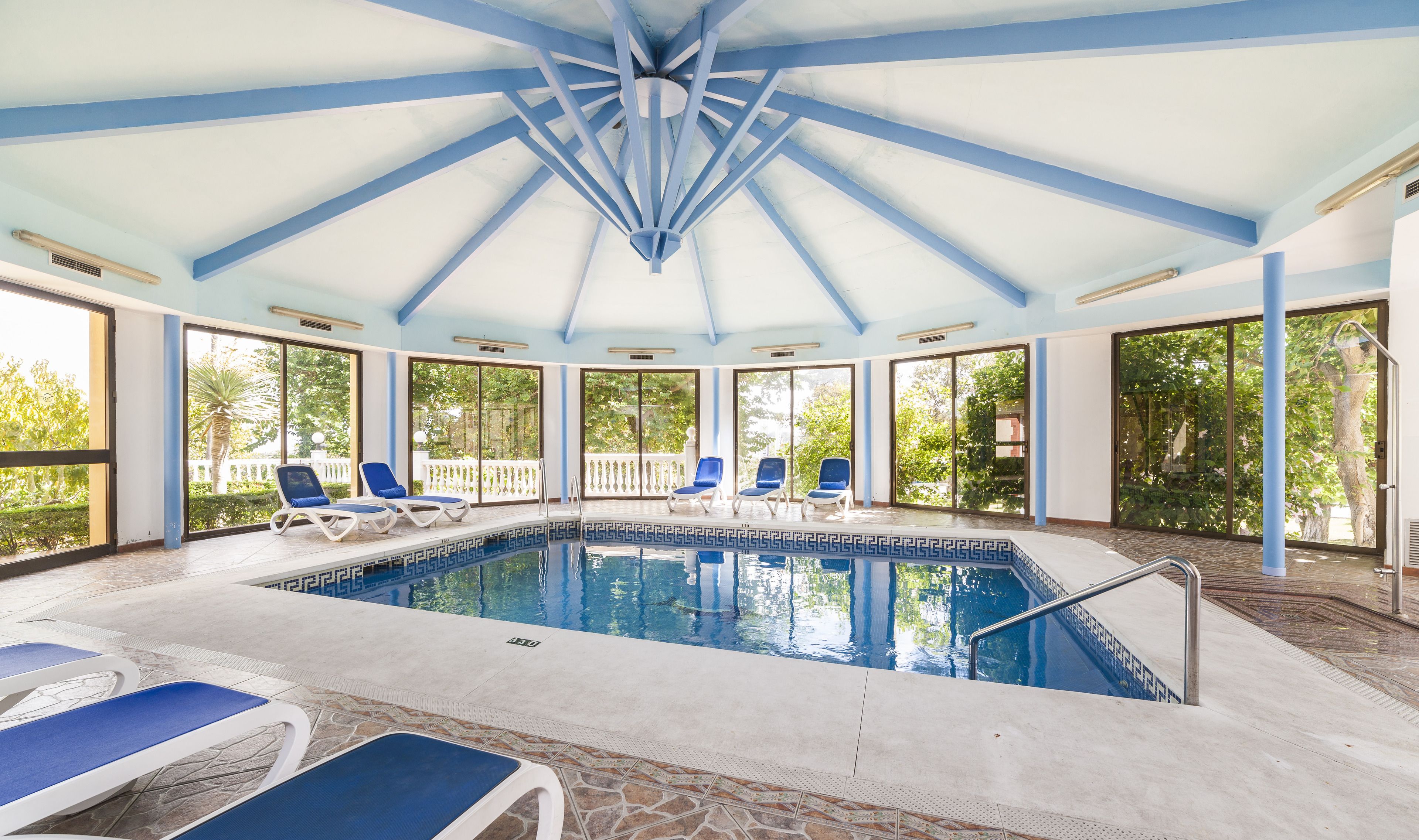 Una piscina interior con vigas de techo azules, ventanas grandes y sillas de descanso.