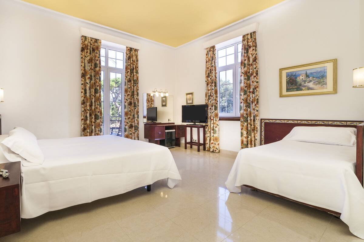 Ein Hotelzimmer mit zwei Betten, einem Schreibtisch mit Fernseher und einem Fenster mit Vorhängen.