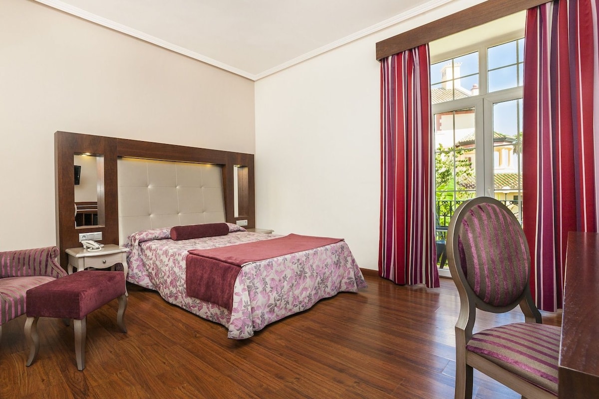 Una habitación de hotel con cama, una silla, un escritorio y una ventana con cortinas.