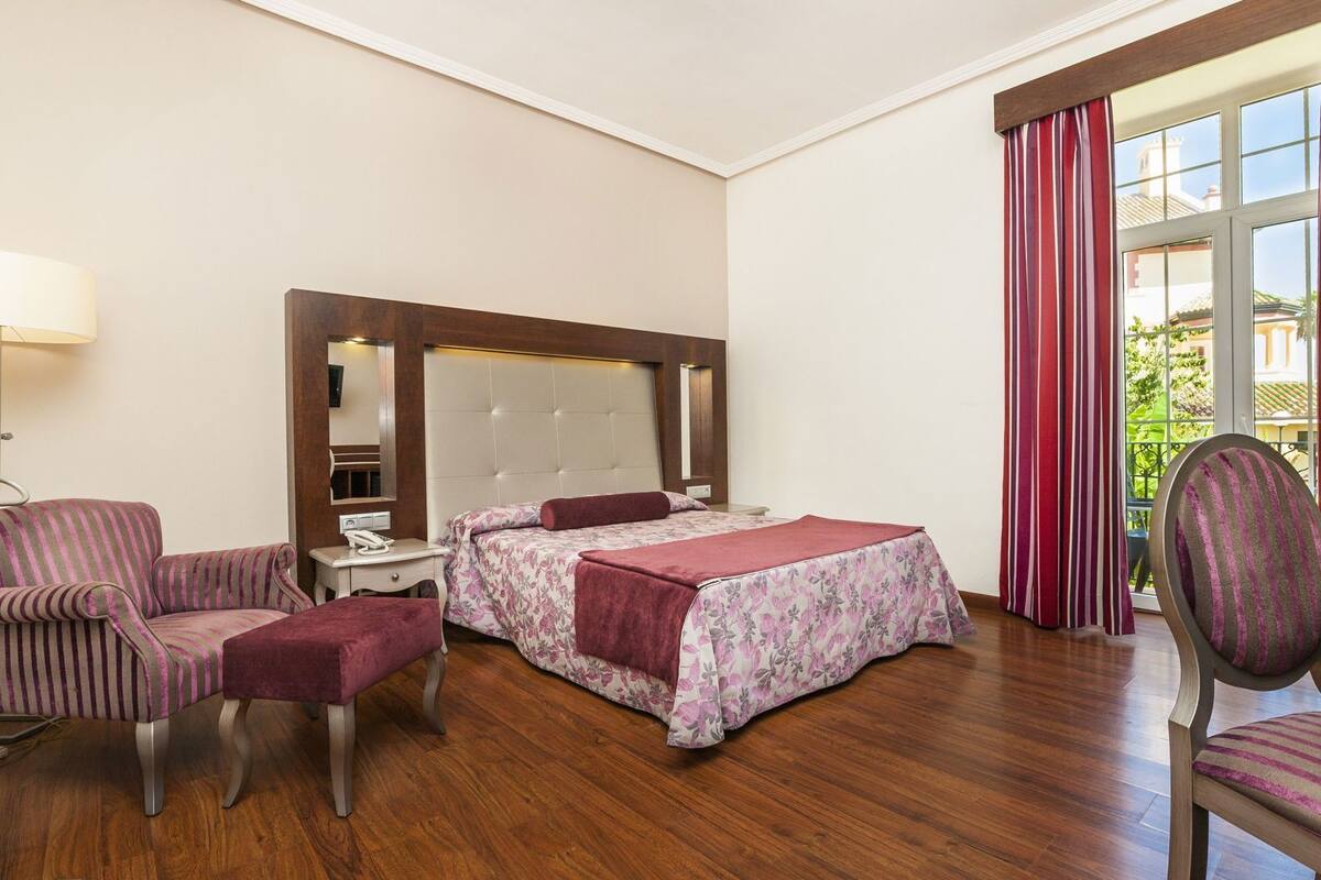 Ein Hotelzimmer mit einem Bett, zwei Sesseln, einem Schreibtisch, einer Lampe und einem fensterbepflanzten Fenster.