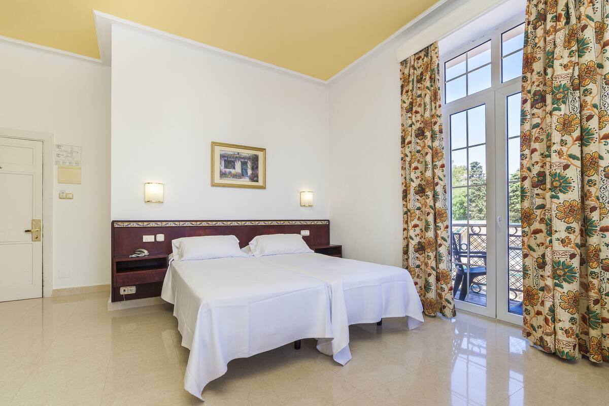 Ein Hotelzimmer mit einem großen Bett, einem Nachttisch, einer Wandlampe, einem gerahmten Bild und einem Fenster mit Vorhängen.