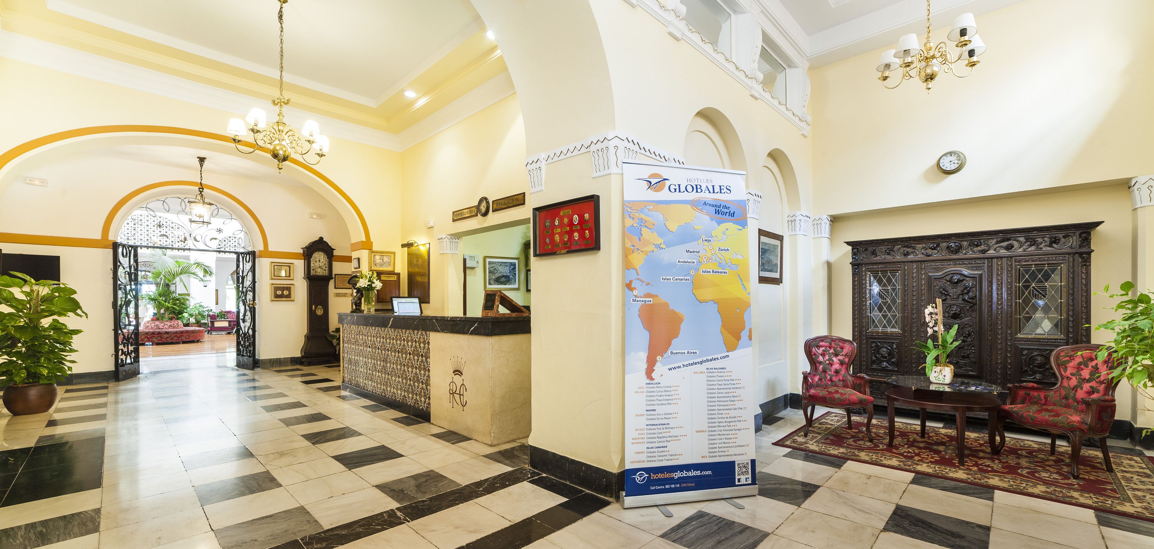 Vestíbulo de hotel con mostrador de recepción, un mapa mundial y decoración clásica, incluyendo un candelabro y sillones tapizados en rojo.