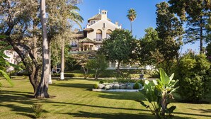 A large, historic building with a dome and multiple windows, surrounded by a well-maintained garden with various trees and a small pond.