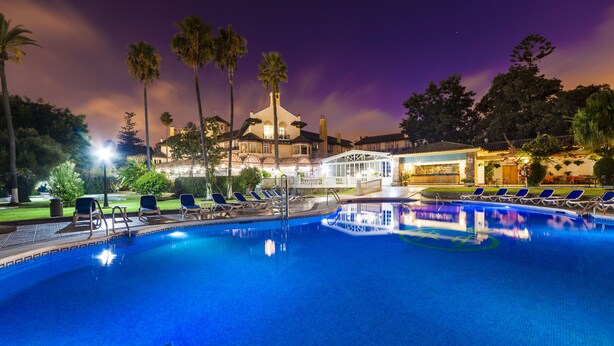 A swimming pool with lounge chairs, palm trees, and a building with a dome roof in the background.
