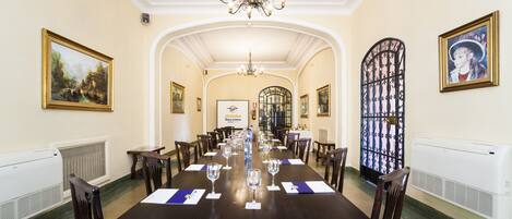 A conference room with a long table set for a meeting, surrounded by chairs, with a chandelier above and framed artwork on the walls.