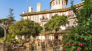 A spacious outdoor patio with multiple tables and chairs, surrounded by lush greenery and a large building with a distinctive architectural style.
