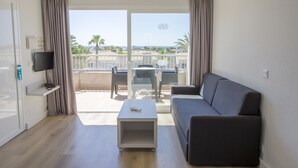 A modern living room with a grey sofa, a small white coffee table, and a TV mounted on the wall. There is a balcony with a table and chairs, and a view of palm trees and a body of water. - Hotel Globales Los Delfines (Ciutadella de Menorca)