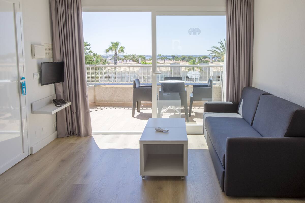 Ein modernes Wohnzimmer mit grauem Sofa, einem kleinen weißen Couchtisch und einem an der Wand befestigten Fernseher. Ein Balkon mit Tisch und Stühlen bietet Blick auf Palmen und ein Gewässer.