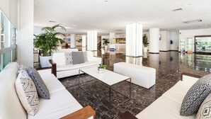 Un vestíbulo amplio con piso de mármol, sofás blancos con cojines estampados y grandes plantas en macetas.