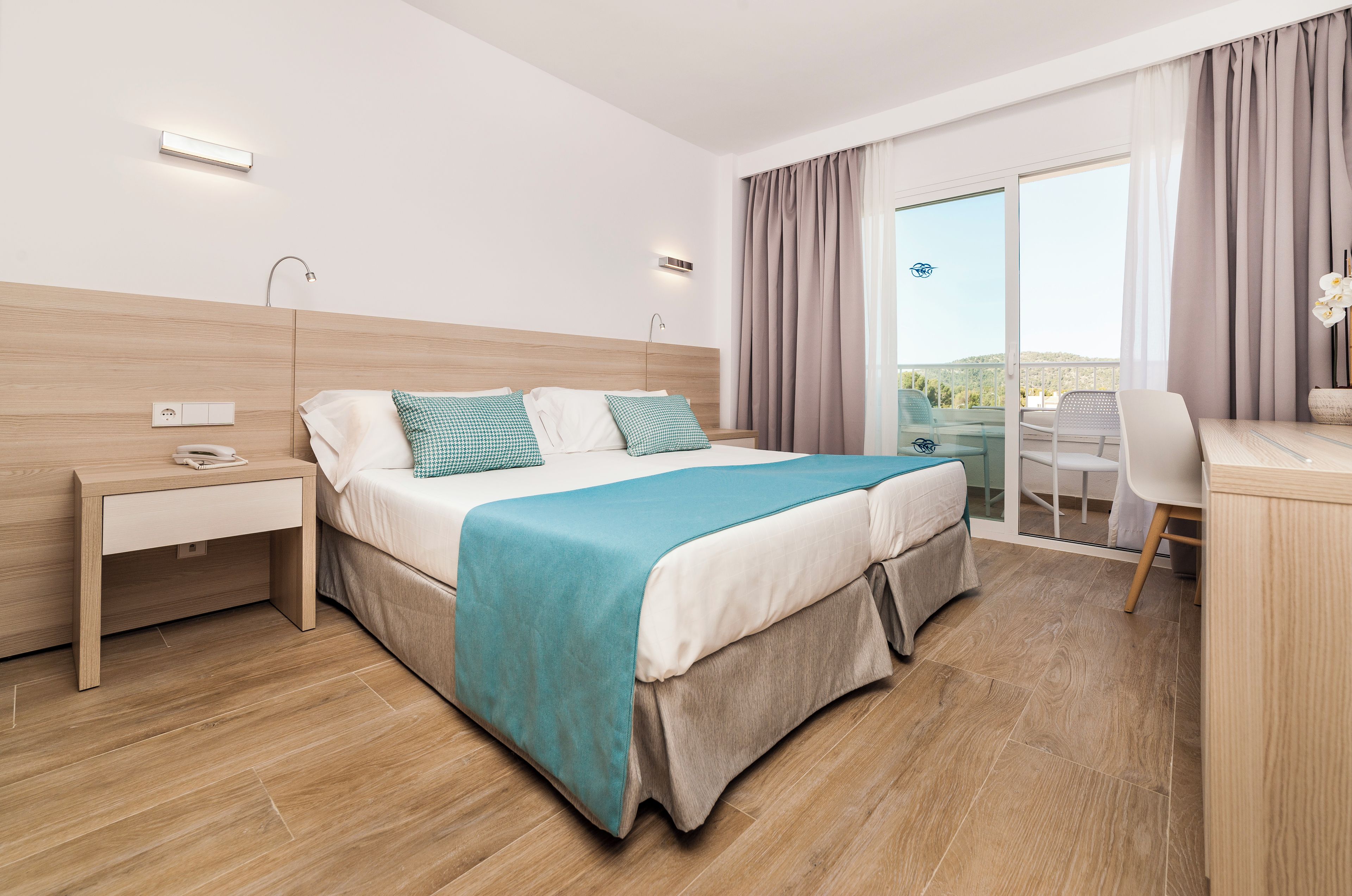 A modern hotel room with a large bed, bedside table, desk, and chair.