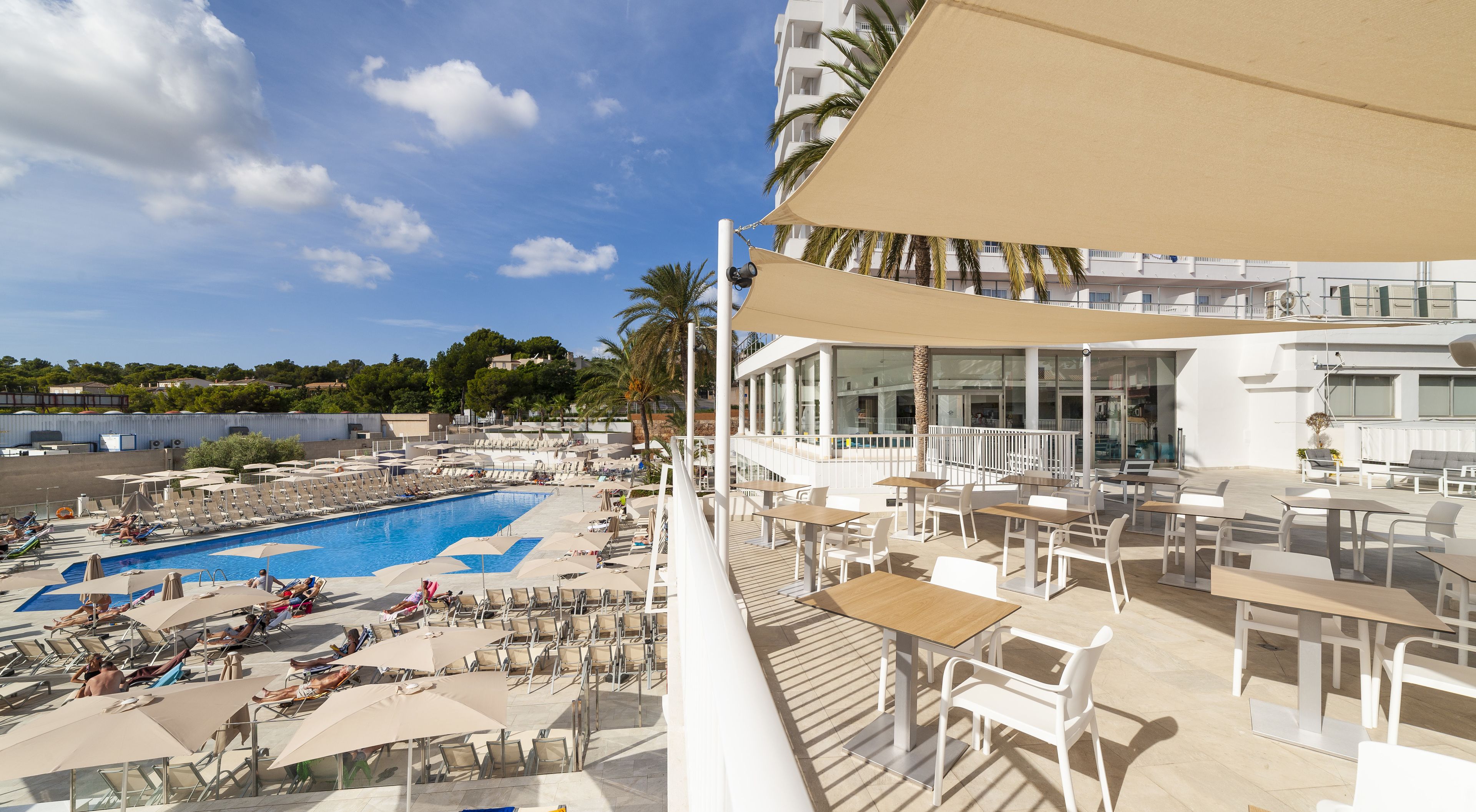 Un área de piscina con sillones de descanso y sombrillas, un comedor con mesas y sillas, y vistas de los edificios y palmeras circundantes.