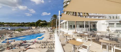 Un área de piscina con sillones de descanso y sombrillas, un comedor con mesas y sillas, y vistas de los edificios y palmeras circundantes.