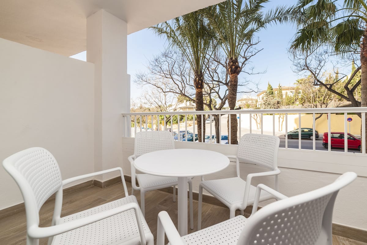 Un balcon avec des chaises blanches et une table, donnant sur une rue bordée de voitures garées et de palmiers.