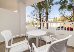 A balcony with white chairs and a table, overlooking a street with parked cars and palm trees. - Globales Mimosa (Calvia)