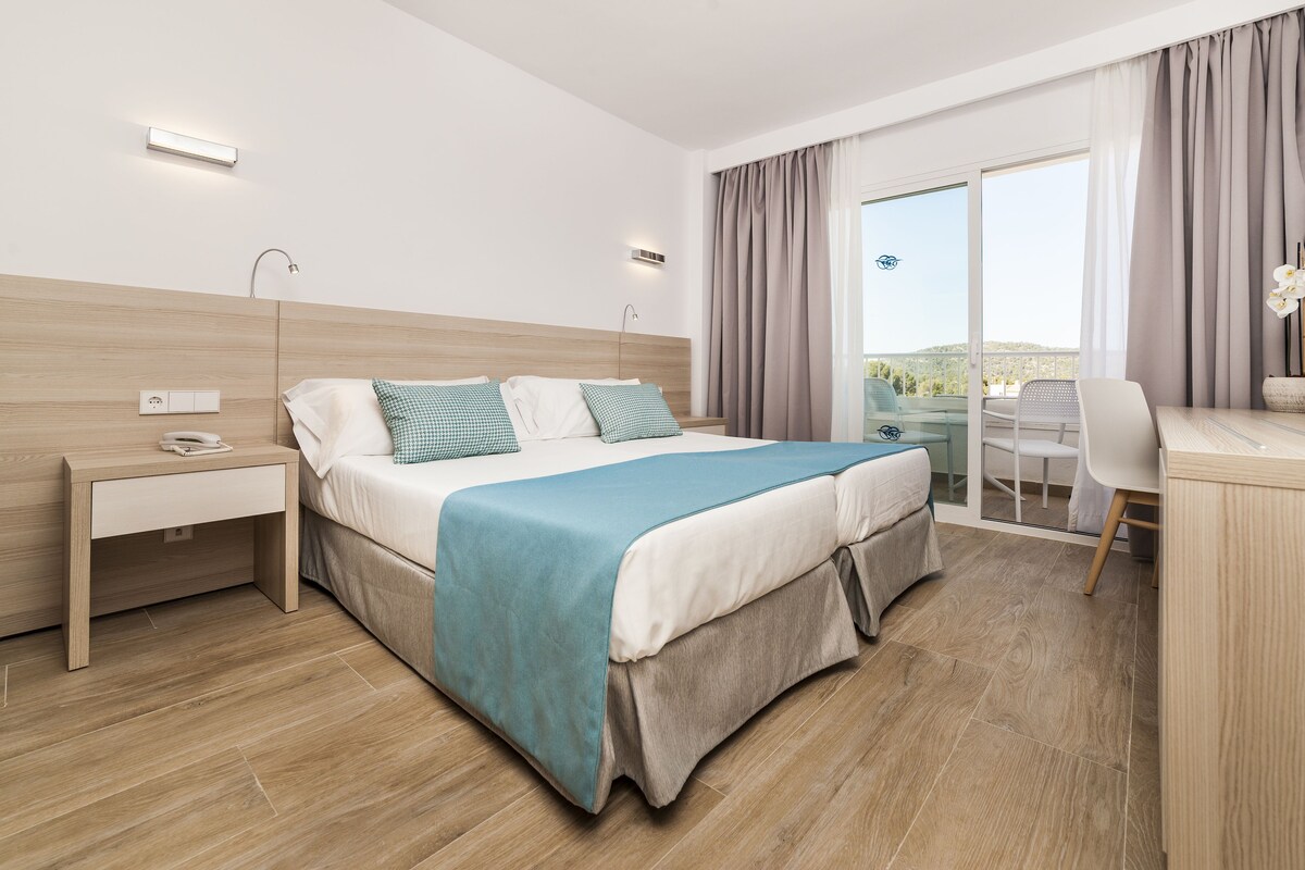 A modern hotel room with a large bed, bedside table, desk, and chair.