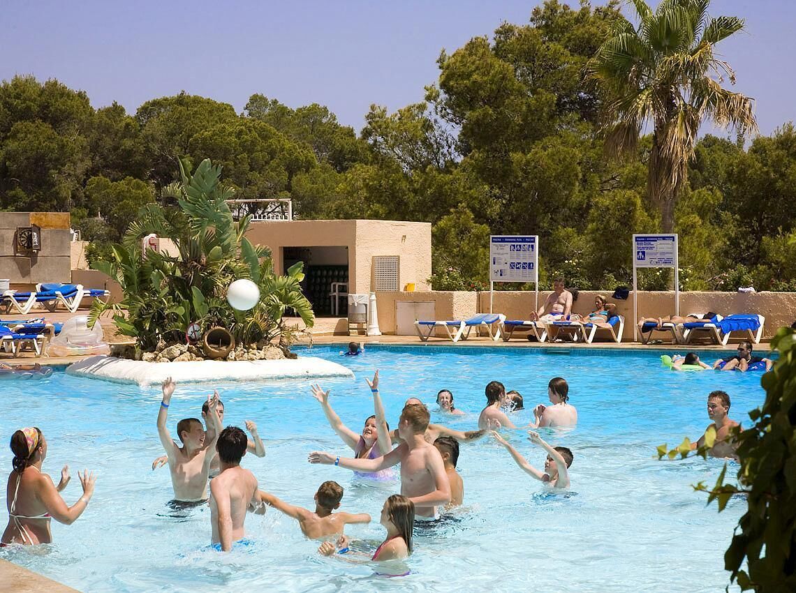 Una piscina con gente disfrutando, rodeada de sillones de descanso y palmeras.
