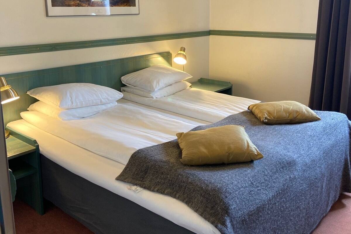 Ein ordentlich bezogenes Bett mit weißen Leinen, einer grauen Decke und zwei Kissen. Darüber an der Wand hängt ein gerahmtes Bild.