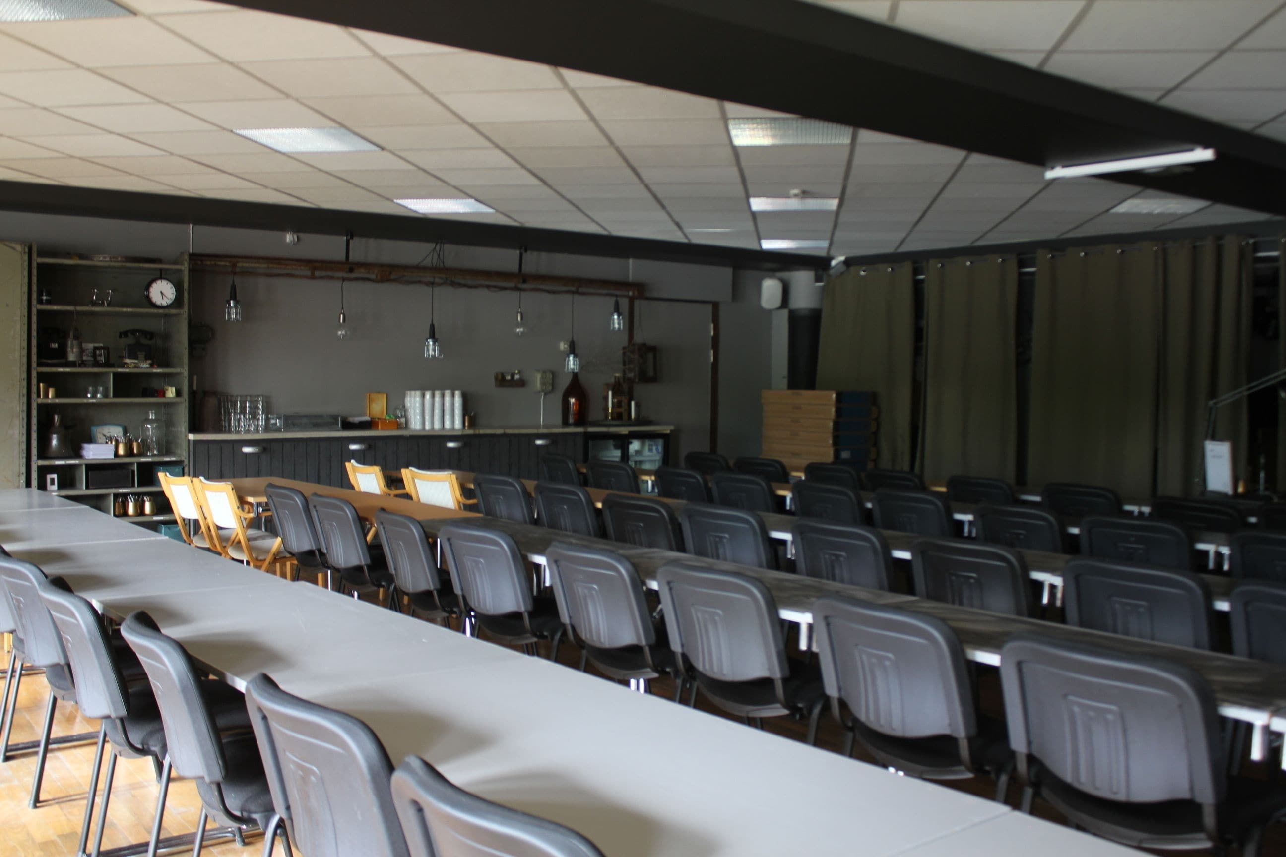 Un salón de conferencias con filas de sillas, una mesa larga y una zona de bar con estantes y botellas.