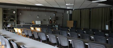 Un salón de conferencias con filas de sillas, una mesa larga y una zona de bar con estantes y botellas.