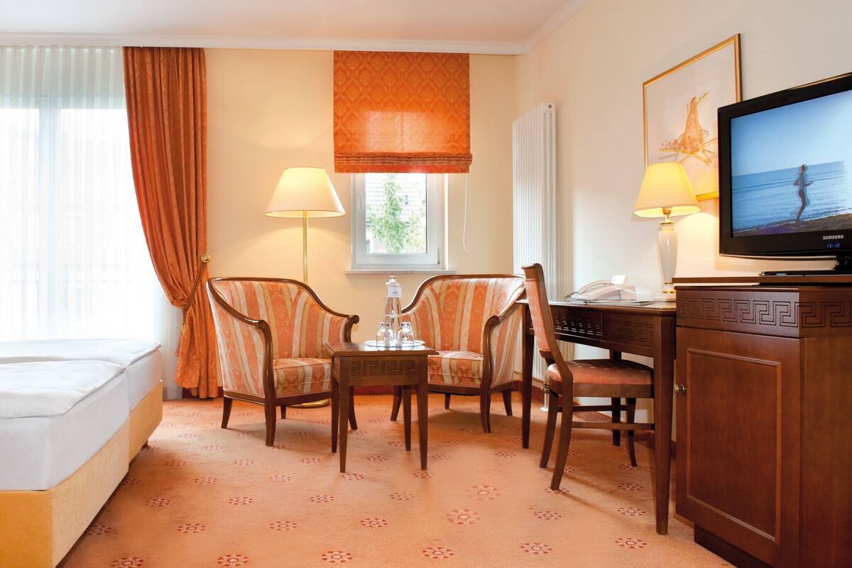 A hotel room with a bed, two chairs, a table, a television, and a window with curtains.