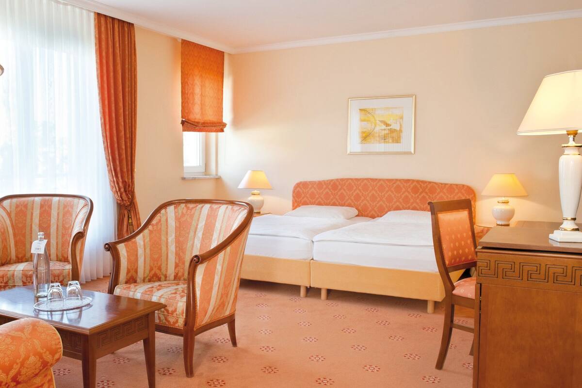 A hotel room with a bed, two armchairs, a small table with a bottle and glasses, a lamp, and a painting on the wall.