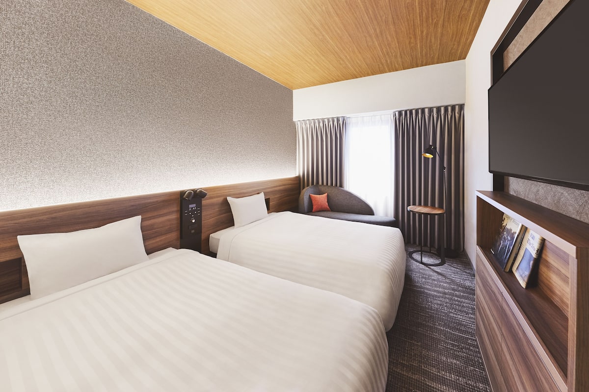 A hotel room with two beds, a wooden headboard, a large window with curtains, a small seating area, and a mounted television.