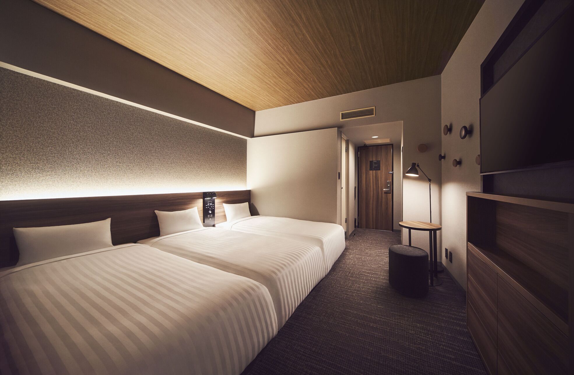 A hotel room with two beds, a wooden ceiling, a textured wall, a small table, and a lamp.