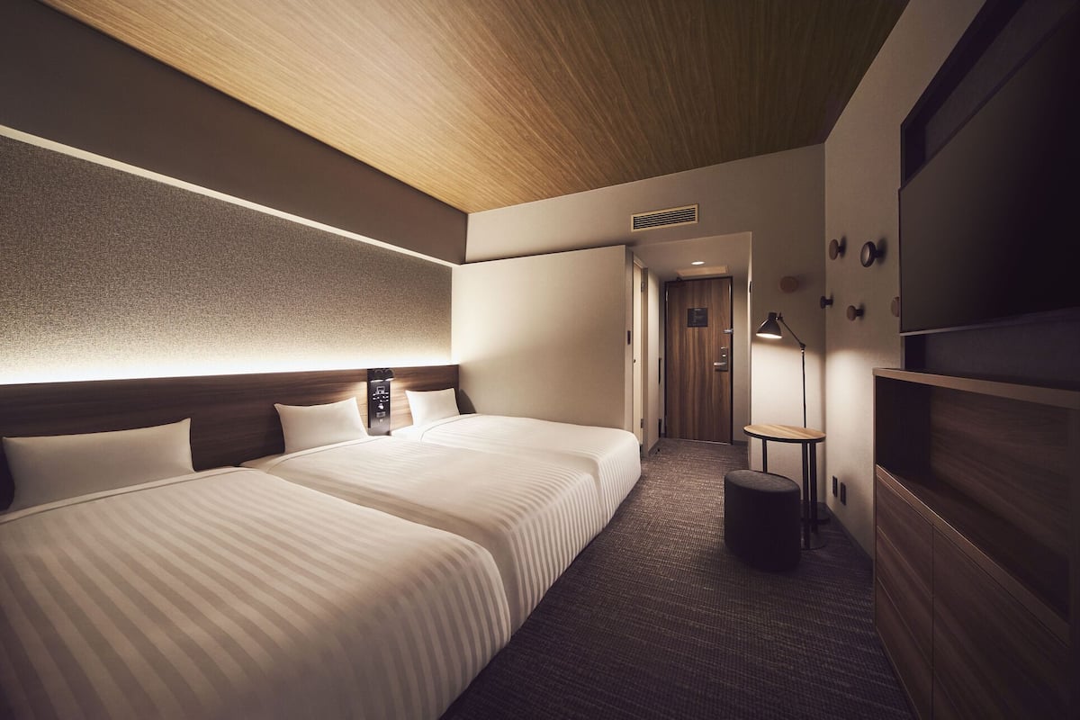 A hotel room with two beds, a wooden ceiling, a textured wall, a small table, and a lamp.