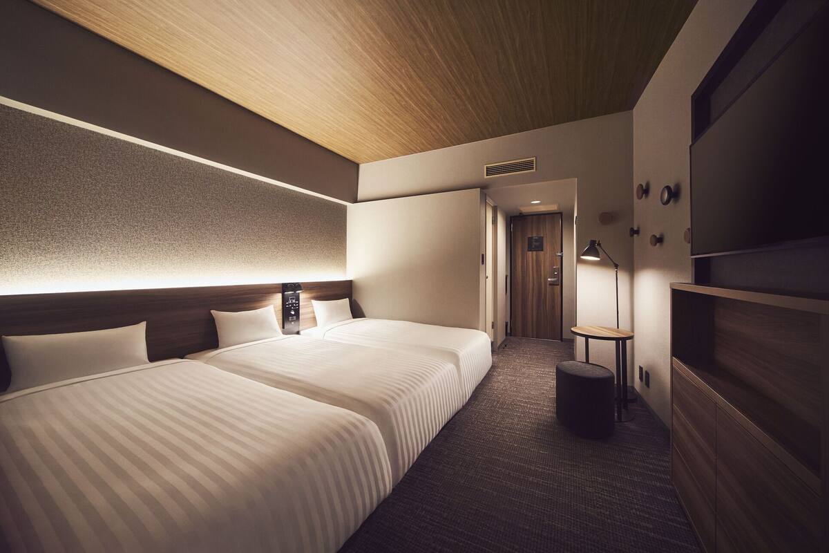 A hotel room with two beds, a wooden ceiling, a textured wall, a small table, and a lamp.
