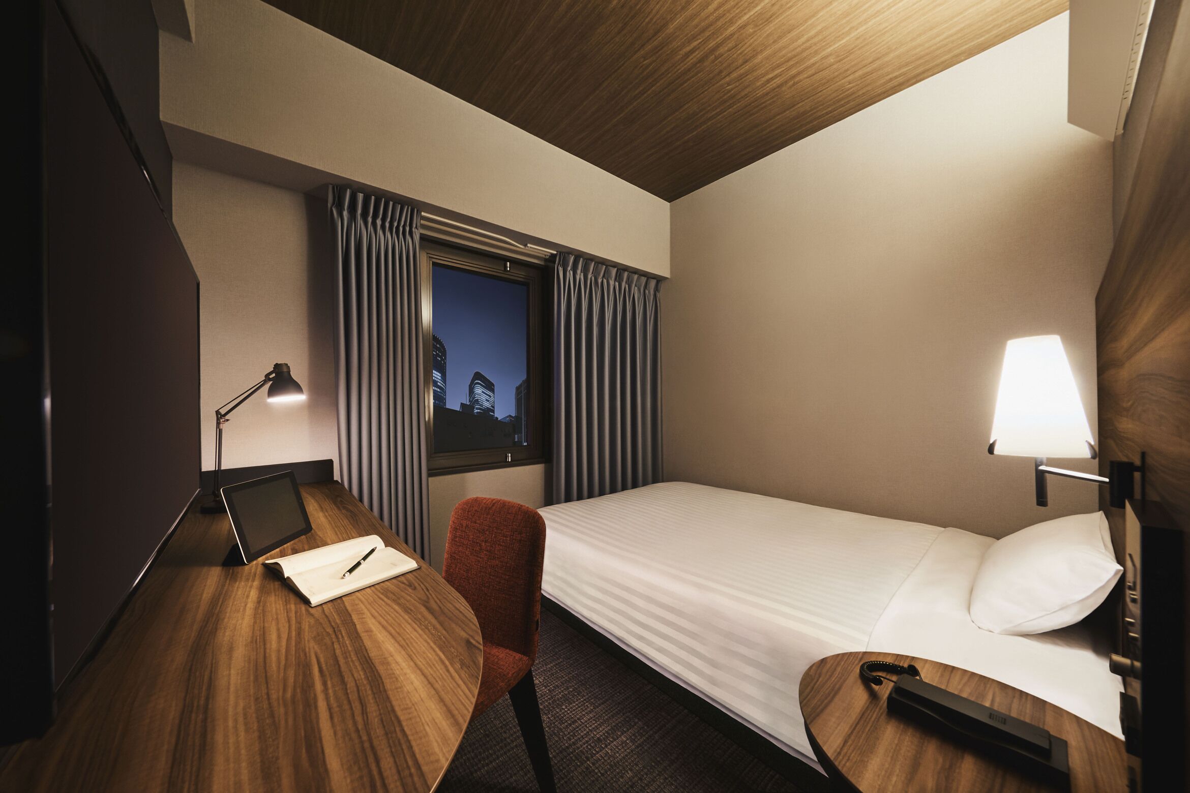 A hotel room with a bed, desk, chair, lamp, and window with curtains.