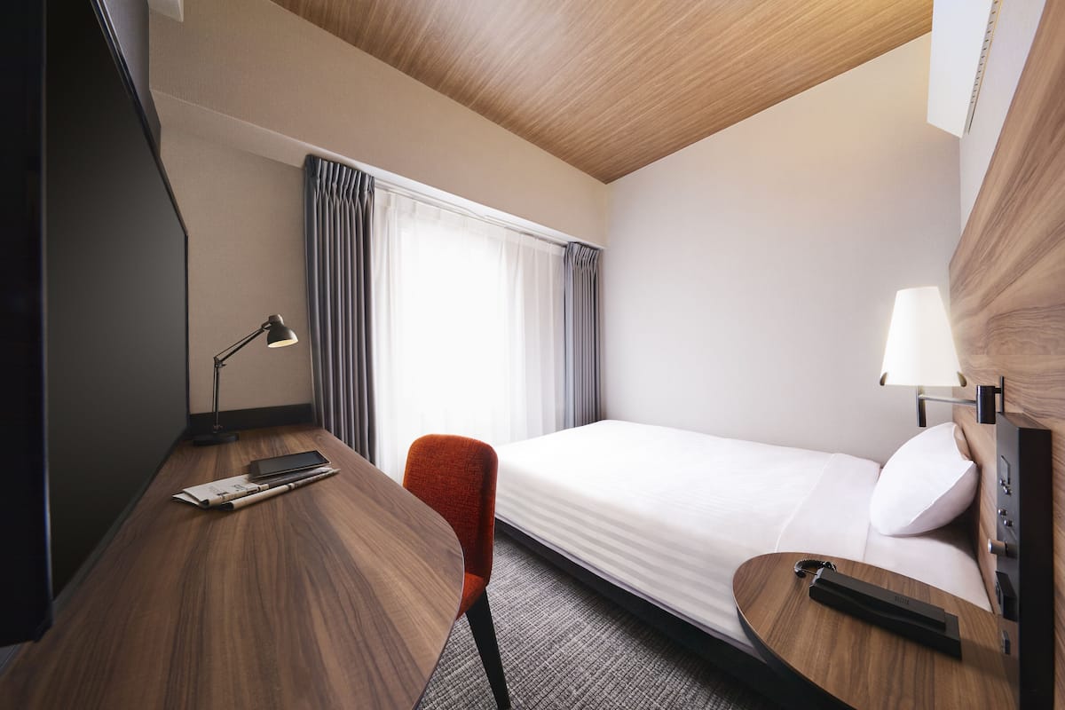 A hotel room with a wooden desk, a red chair, a bed with white linens, a lamp, and a window with curtains.