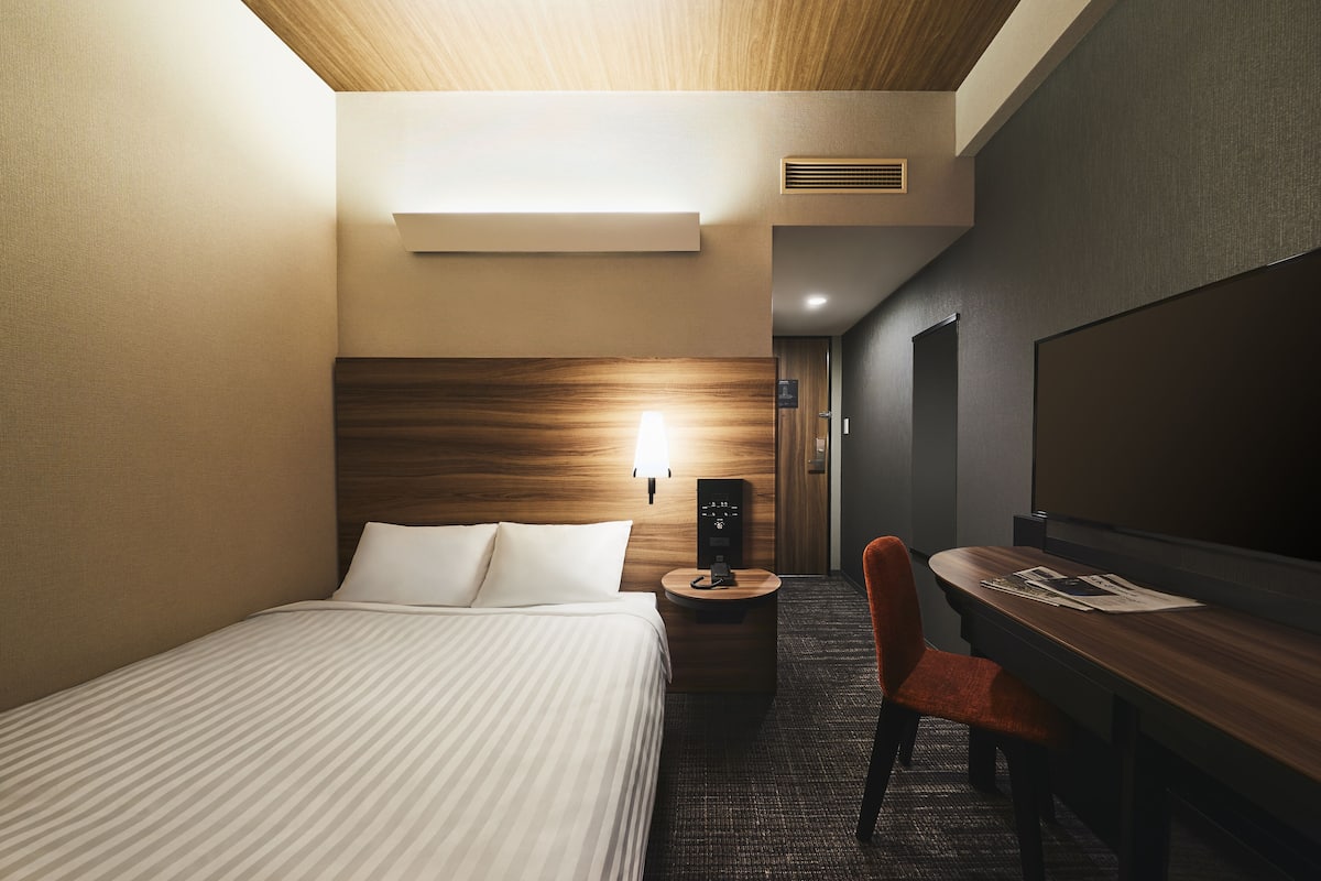 A hotel room with a bed, a desk, a chair, a lamp, and a television.