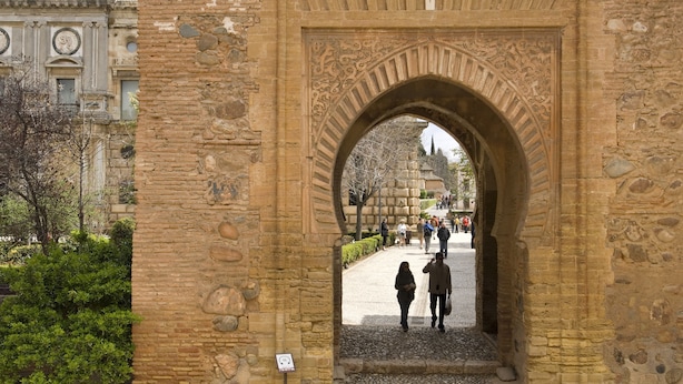 An ancient stone archway with intricate carvings, leading to a paved area with people walking.