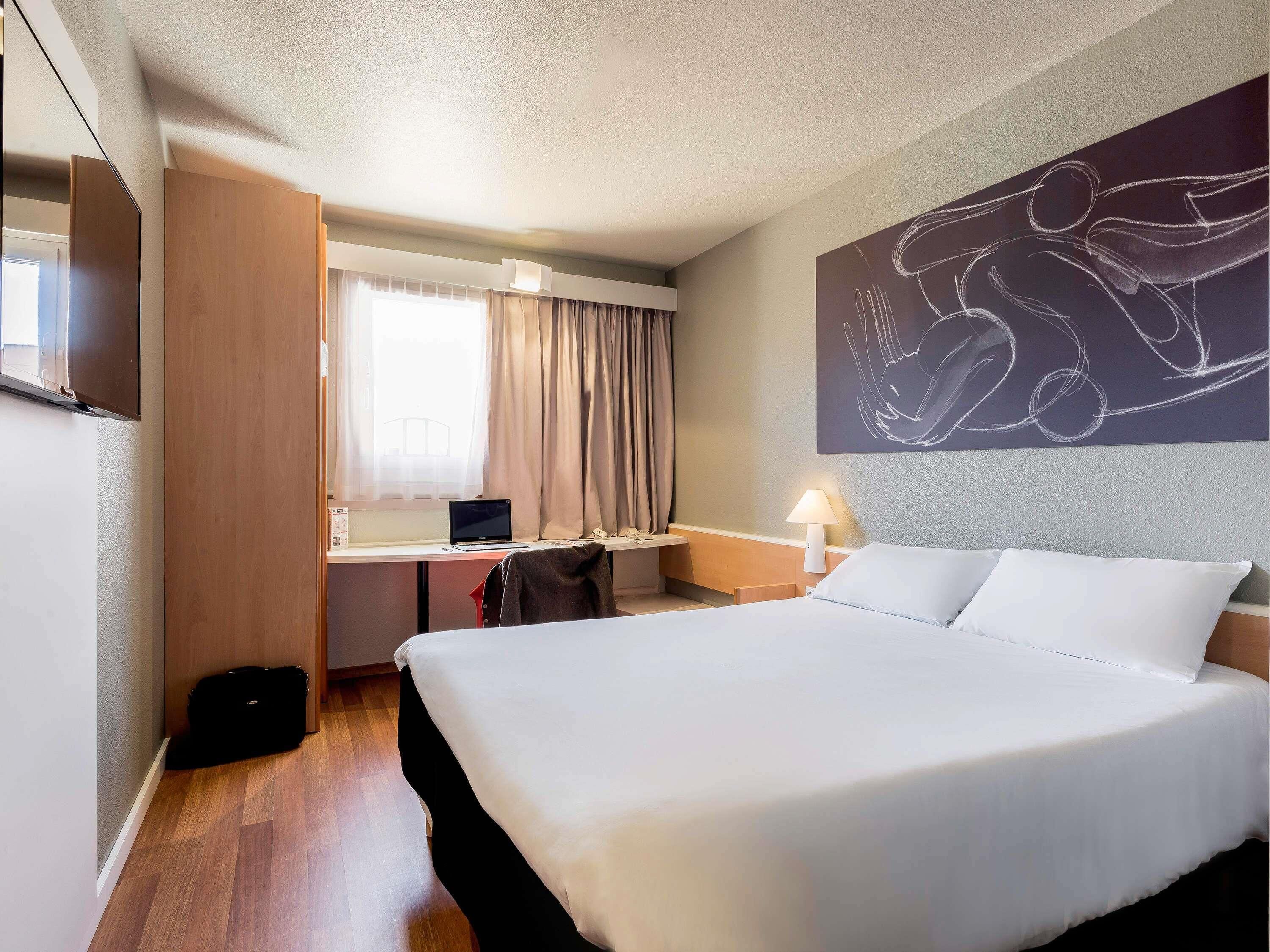 A hotel room with a bed, desk, laptop, and a wall art piece.