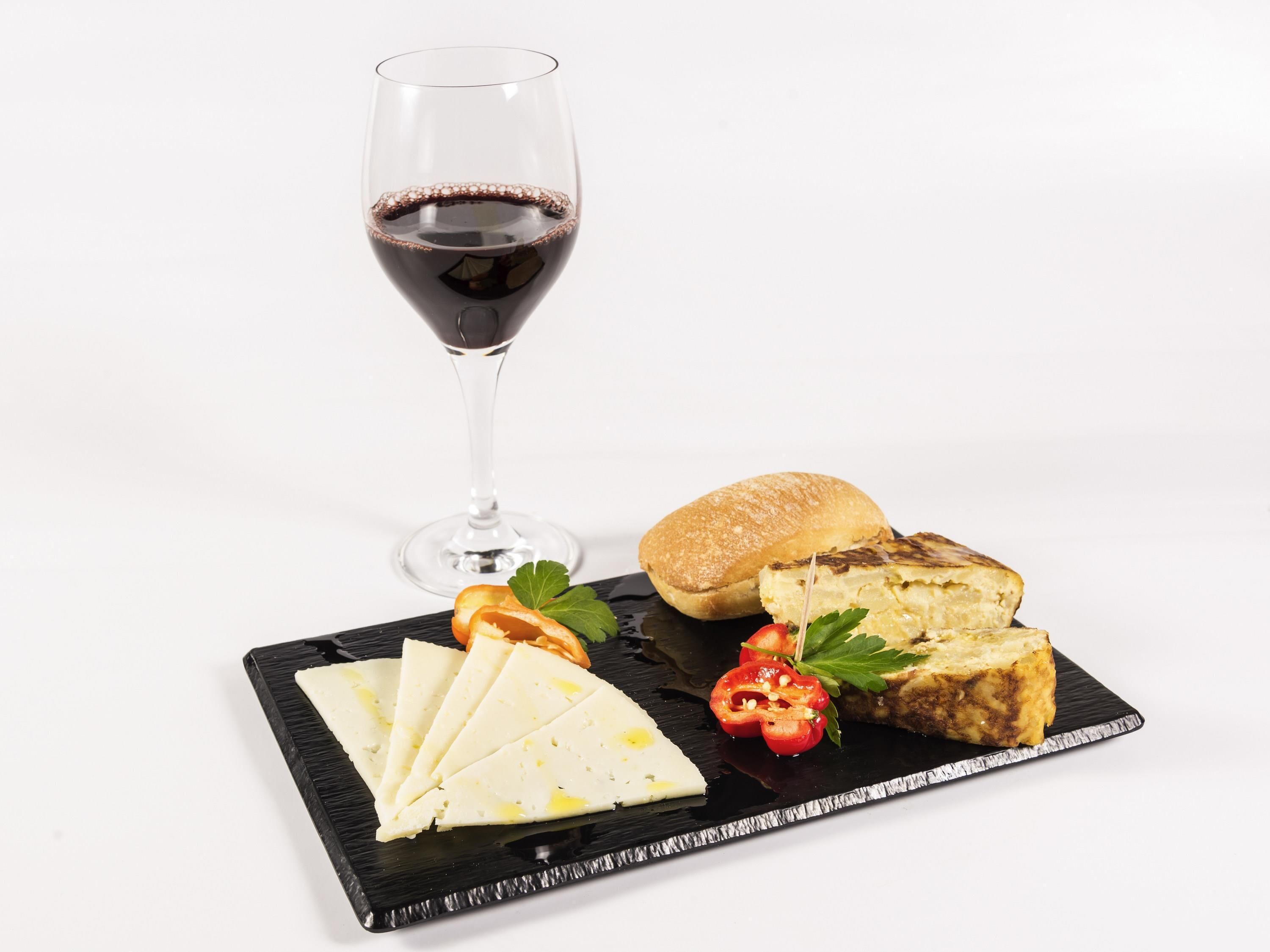 A glass of red wine, a baguette, cheese, and a small salad on a black slate plate.