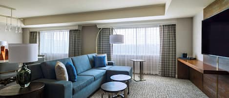 A modern hotel room with a sectional sofa, two small round tables, a flat-screen TV, and a large window with curtains.