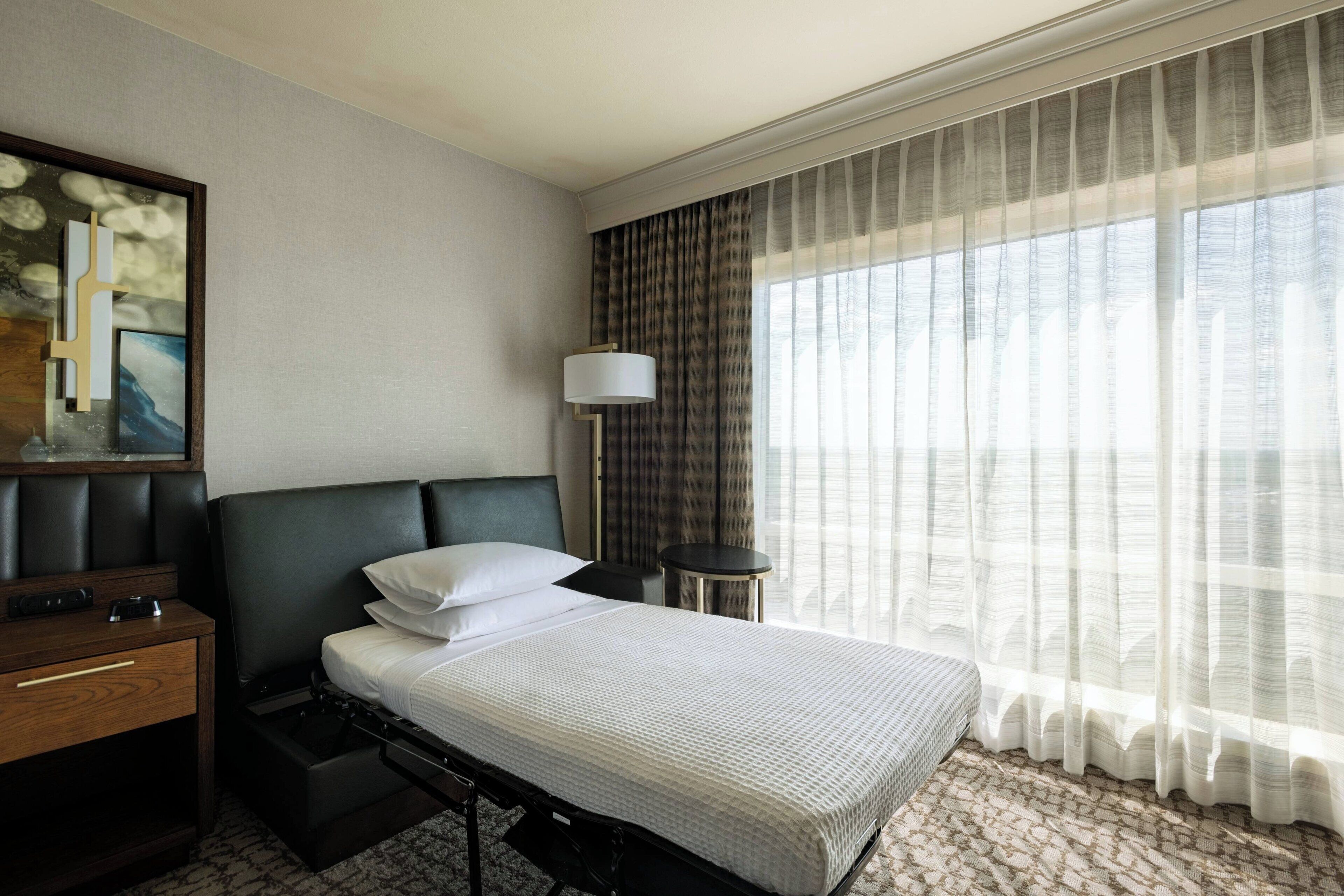 A hotel room with a bed, a bedside table, a lamp, and a large window with curtains.