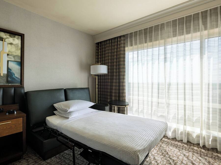 A hotel room with a bed, a bedside table, a lamp, and a large window with curtains.